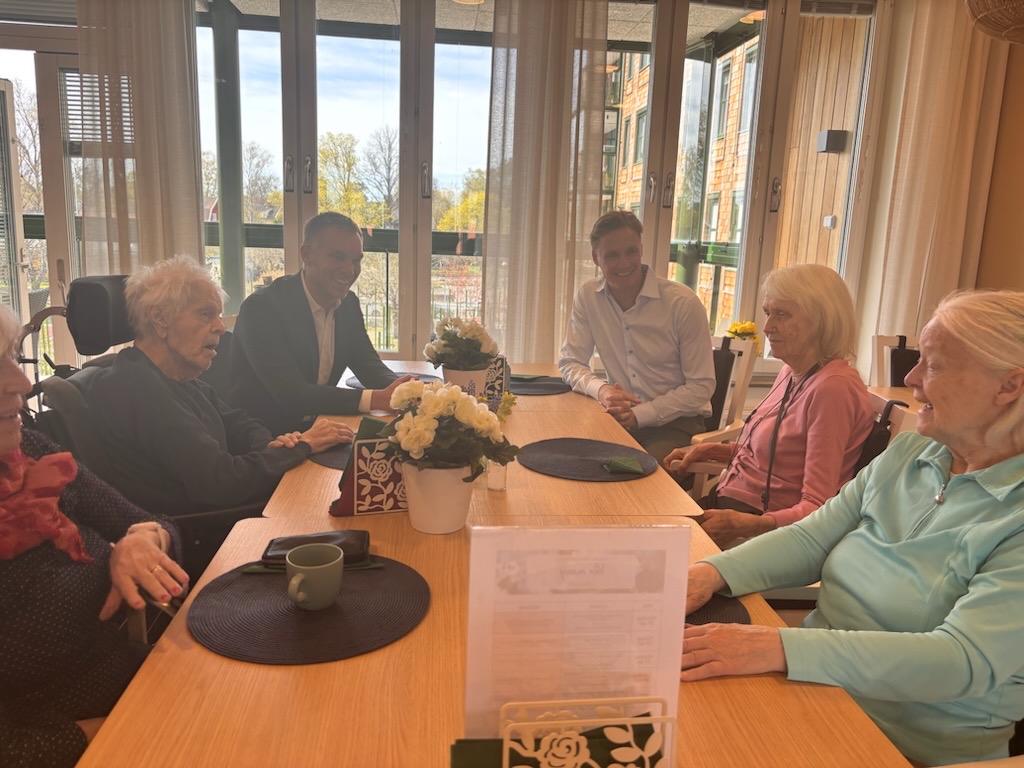 Civilminister Erik Slottner och Attendokoncernens VD Martin Tivéus sitter vid ett bord och samtalar med boende på Attendo Näsbyterrassen.