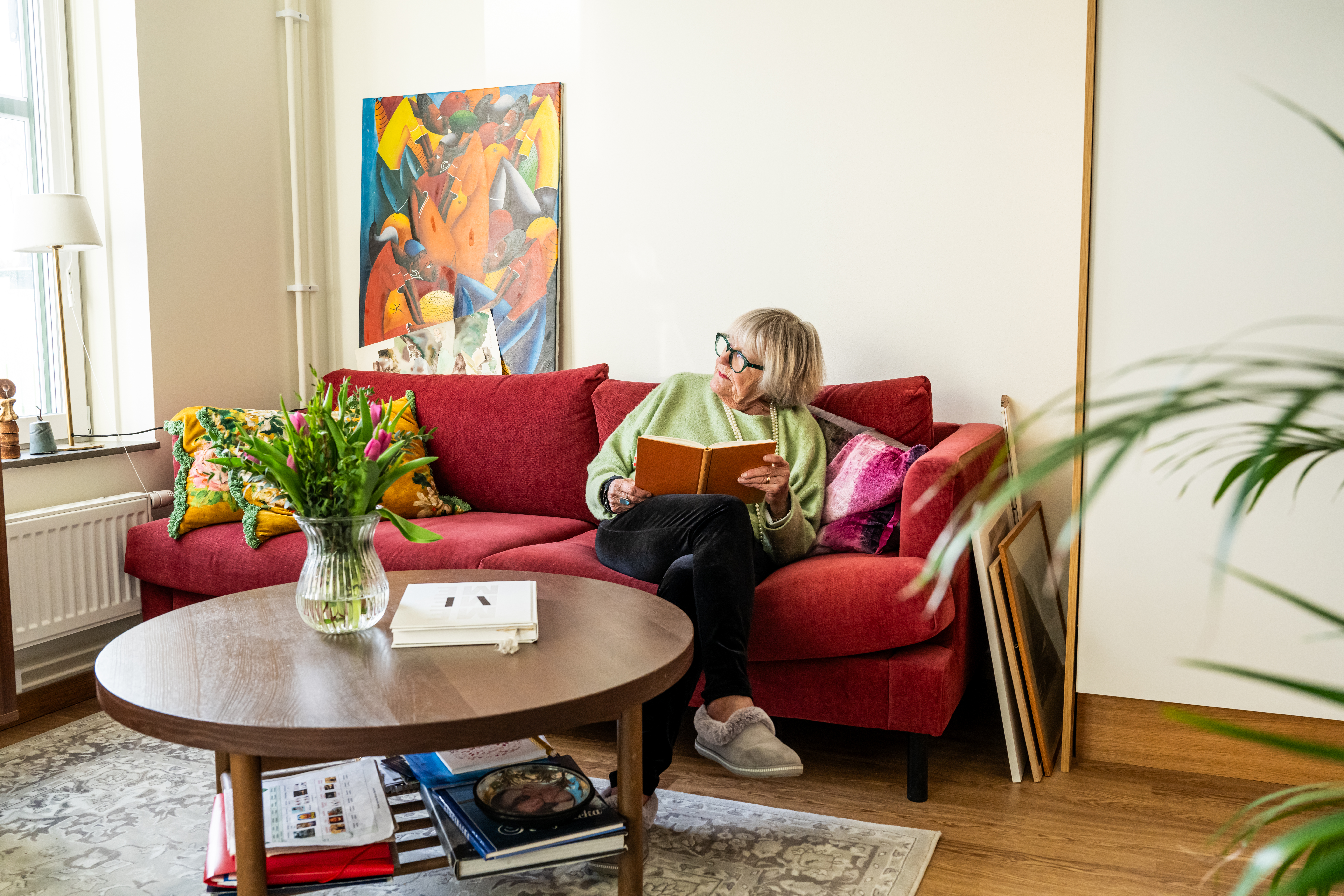 Äldre kvinna sitter i soffa i sin lägenhet på äldreboende med en bok i handen och ser drömmande ut genom fönstret.