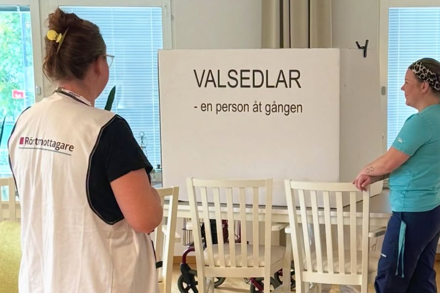 En rullator skymtar under ett valbås, röstmottagare och Attendos personal väntar vid sidan av