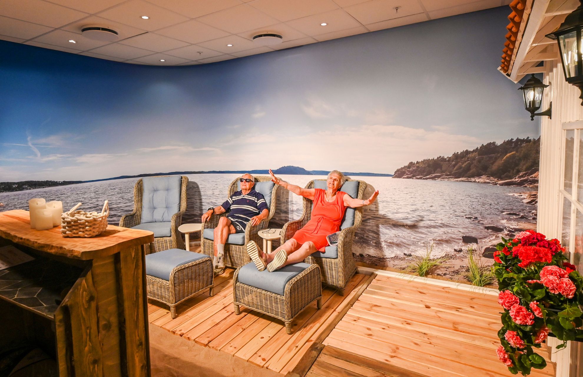 Ett par sitter i solstolar med en drink i handen i Attendo Lundbyparks solrum. I bakgrunden är en tapet över hela väggen som visar hav och stenar.