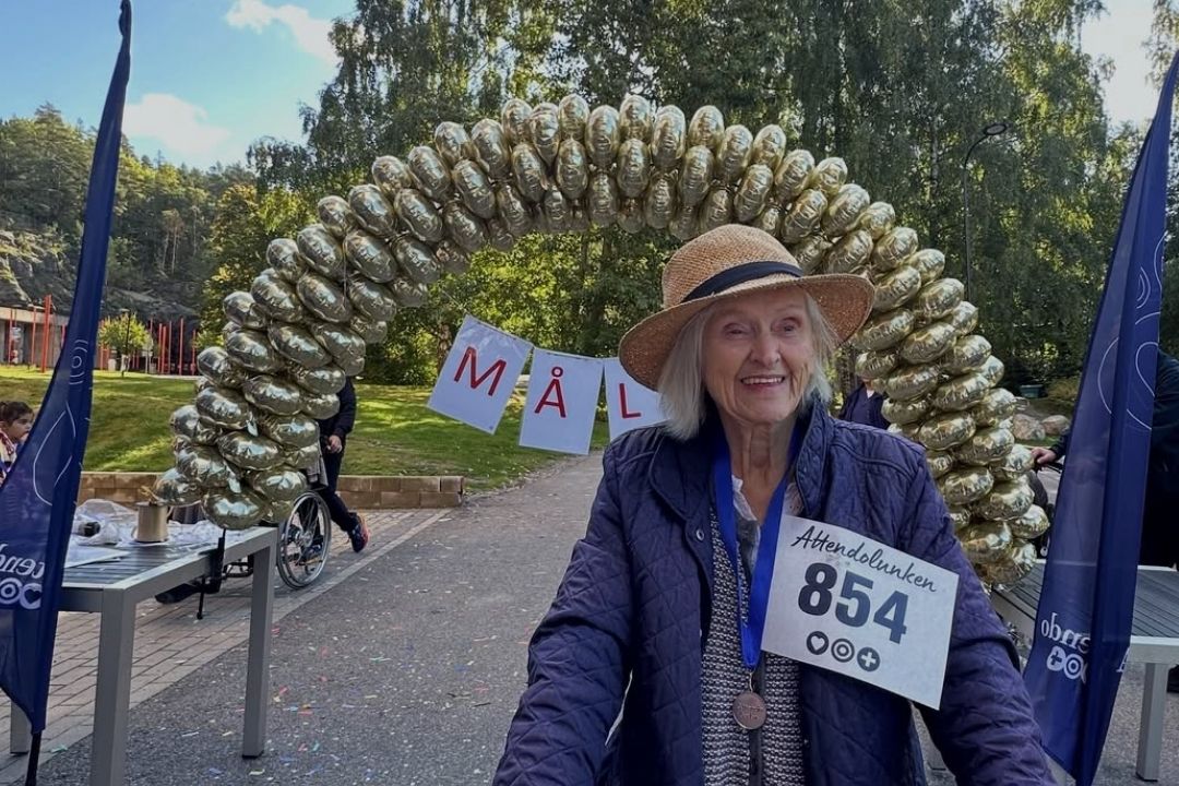 Äldre kvinna med nummerlapp och rullator syns framför målgången och en enorm ballongbåge