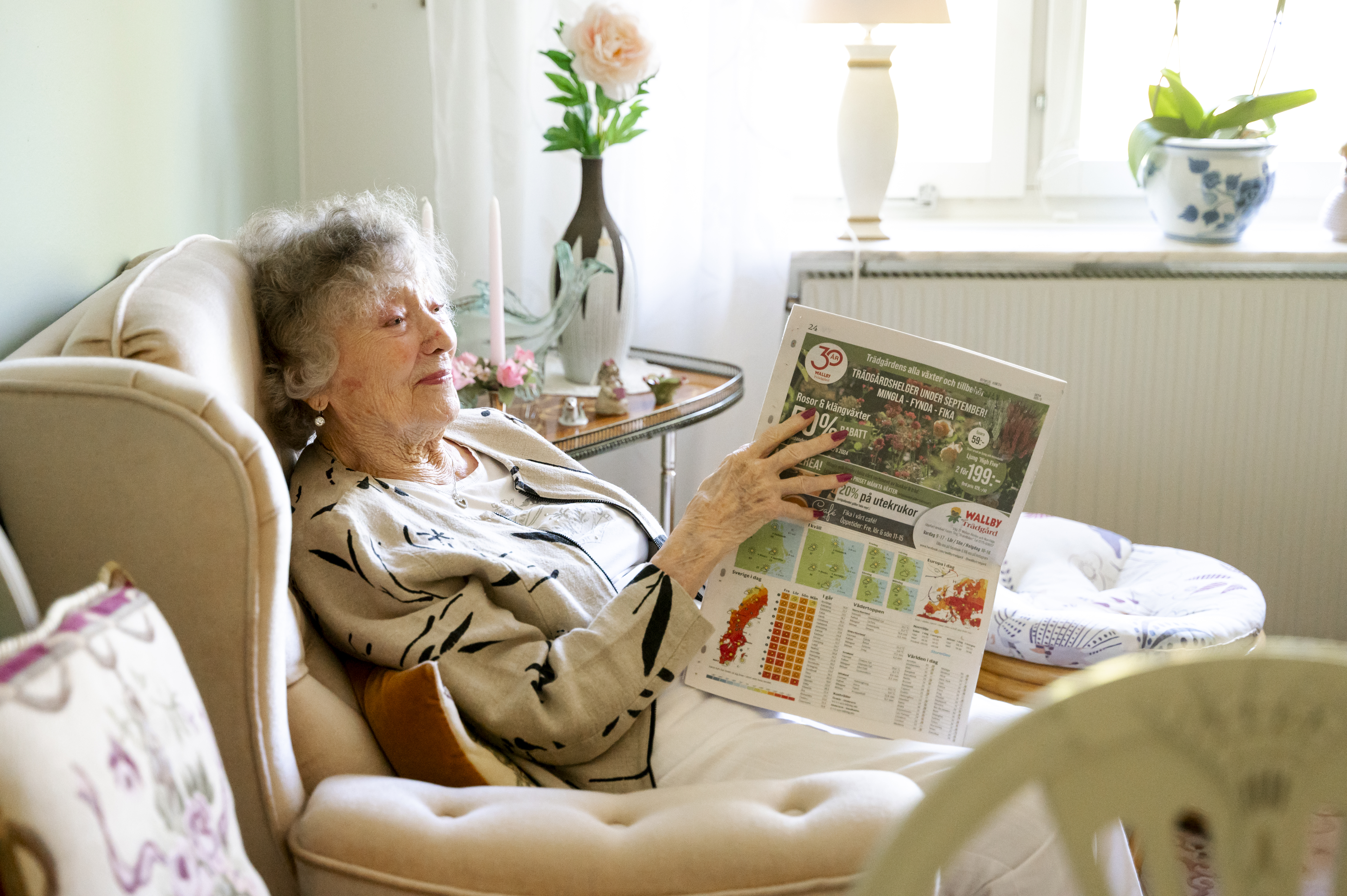 Äldre kvinna sitter i en fåtölj och läser en tidning i ett ljus inrett rum med blommor och dekorativa föremål
