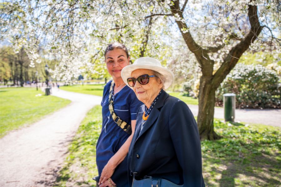 Personal från hemtjänst och äldre kvinna på promenad i vacker park med blommande träd